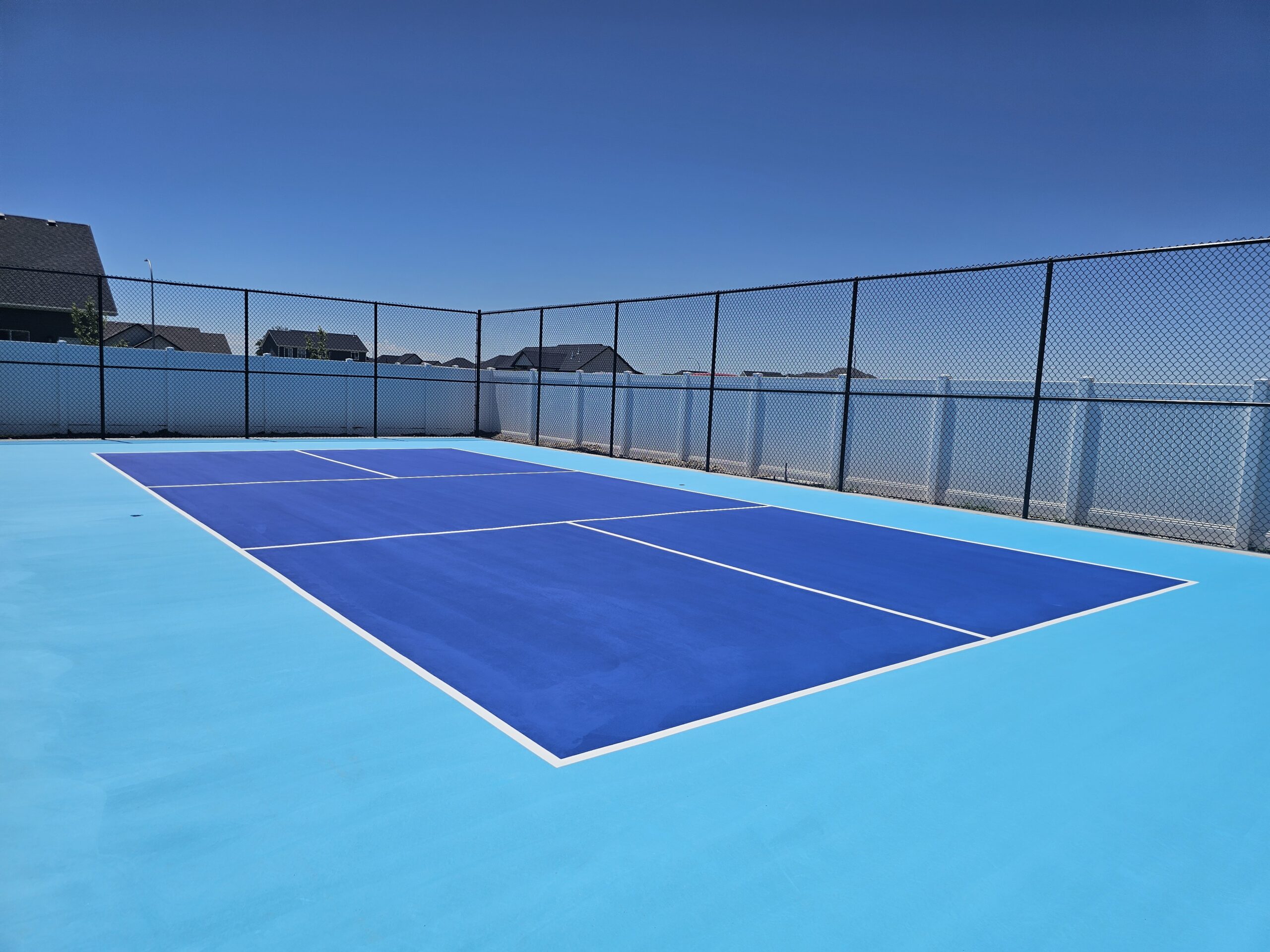 A newly installed pickleball court, built from the ground up. The court is light blue and Kansas City Blue with black vinyl fencing and sports court lighting.